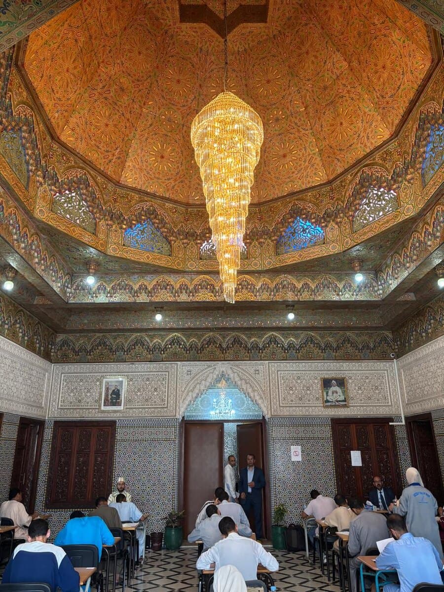 Vue d'ensemble de la salle : plafond peint, lustre et rangées de pupitres.